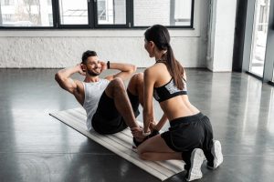 Sporty girl helping happy young man doing abs exercise on yoga mat in gym