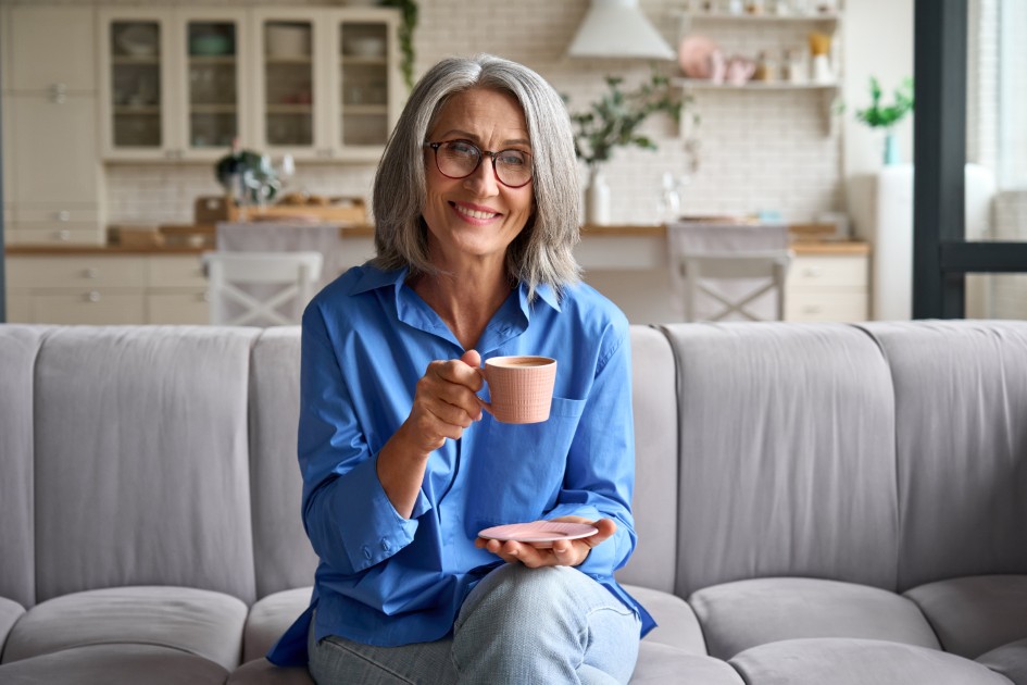 Senior-smiling-60s-woman-portrait-indoors-looking-at-camera-with-cup-of-tea-coffee