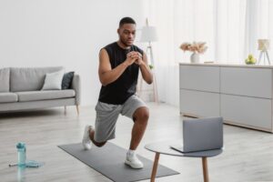 African-Man-Training-At-Laptop-Doing-Forward-Lunge-At-Home