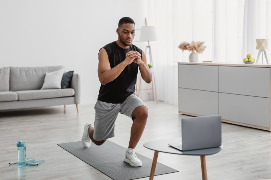 African-Man-Training-At-Laptop-Doing-Forward-Lunge-At-Home