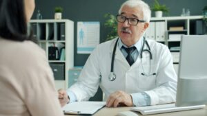 Friendly senior doctor is talking to female patient during appointment in clinic discussing disease and treatment. Communication and medicine concept
