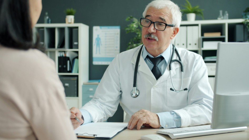 Friendly senior doctor is talking to female patient during appointment in clinic discussing disease and treatment. Communication and medicine concept